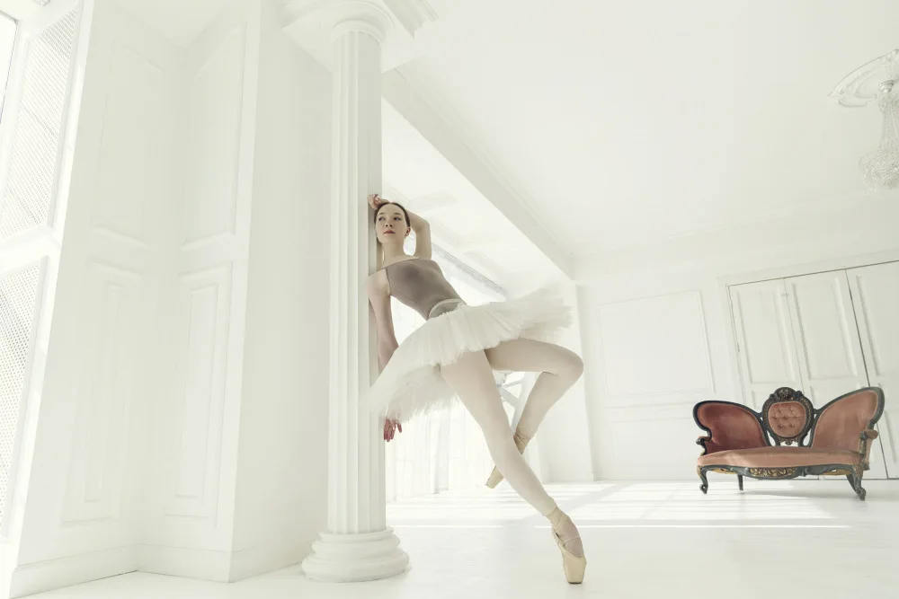 Alexandr: ballerina in a light bodysuit and tutu is standing in the room leaning on a pole
