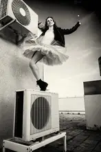 Alexandr: a ballerina in tutu, jacket and boots climbed on the air conditioner and poses against the sky